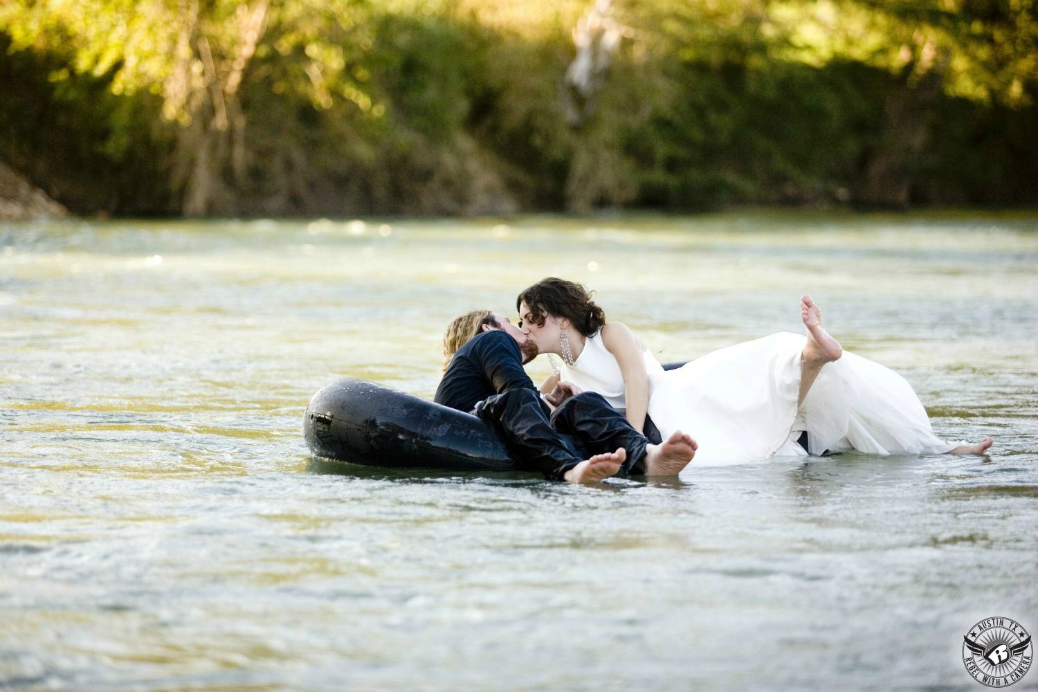 Dark haired girl wears mother wedding dress barefoot floating down the river in a tube kissing a blond guy wearing a tux shirt and black pants also barefoot and also in an inner tube on the Guadalupe River with green trees in the background in this lighthearted summer engagement picture near Bastrop  
